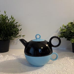 Könnte beinhalten: Zweifarbiges Tee-Set. Die Teekanne ist oben schwarz mit einem hellblauen Deckel und Ausguss. Die untere Hälfte ist hellblau und bildet die Tasse. Der Griff ist ein schwarzer Kreis. Das Set steht auf einer weißen Spitzendecke.