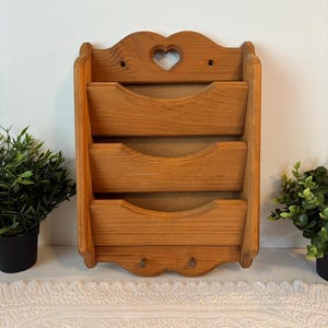 Puede incluir: Organizador de pared de madera con tres compartimentos en niveles. La parte superior presenta un recorte en forma de corazón. El organizador es de madera clara y tiene una base festoneada con dos pequeños ganchos.