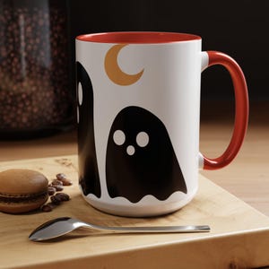Op de afbeelding: Witte keramische mok met een rode handgreep en binnenkant. De mok heeft een zwart spookontwerp met een halve maan. Een lepel en een macaron liggen op een houten snijplank.