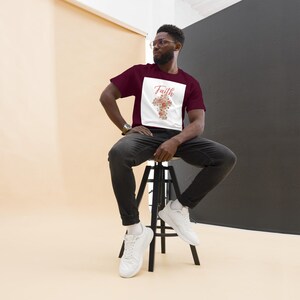 Peut inclure: T-shirt bordeaux avec un rectangle blanc portant le mot "Faith" et une croix florale. La personne porte un jean noir et des baskets blanches. Le fond est un mélange de beige et de noir.