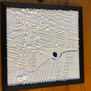 May include: A framed 3D relief map of a city, featuring white buildings and streets against a white background. The map is enclosed in a black frame and mounted on a wooden surface. The map includes a lake and a river.