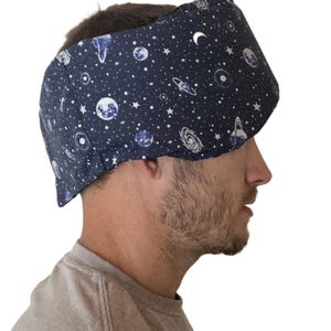 May include: A dark blue sleep mask featuring a celestial pattern of white stars, planets, and crescent moons. The mask is made of fabric and designed to block out light. The mask is worn over the eyes and forehead.