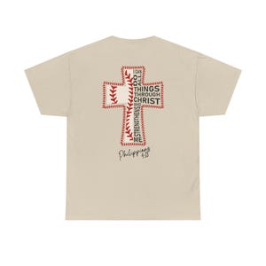 Puede incluir: Una camiseta beige con un diseño de cruz con temática de béisbol en la espalda. La cruz está hecha de costuras de béisbol y una pelota de béisbol. El texto dentro de la cruz dice "Todo lo puedo en Cristo que me fortalece" y "Filipenses 4:13".