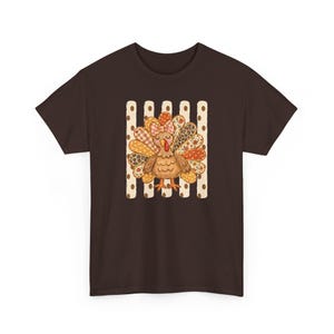 Peut inclure: Un t-shirt à manches courtes marron foncé avec un motif central représentant une dinde à la queue colorée et à motifs. La dinde porte un petit nœud sur la tête et se trouve devant un fond à rayures verticales crème avec des pois marron.