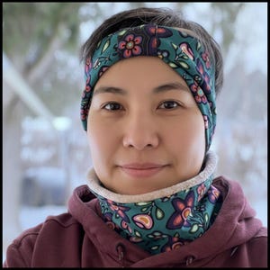 Op de afbeelding: Een turquoise hoofdband en nekwarmer set met een bloemenpatroon in roze, paars en wit. De hoofdband wordt op het hoofd gedragen en de nekwarmer om de nek. De persoon draagt een bordeauxrode hoodie.