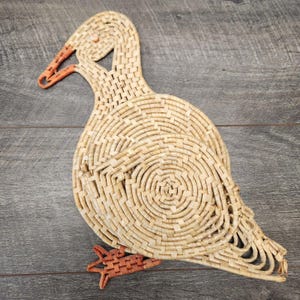 May include: A woven duck-shaped trivet in natural and orange tones. The duck's body features a spiral design, with an orange beak and feet. This decorative item is made of woven material.