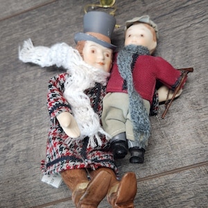 May include: Two porcelain dolls with painted features. One doll wears a gray top hat, a white knit scarf, and a red, black, and white houndstooth coat. The second doll wears a gray cap, a gray knit scarf, a red jacket, and beige trousers.