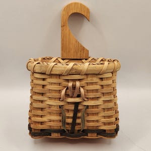 May include: A woven basket with a light tan finish and a flat wooden hook attached to the top. The basket features a square shape with a decorative tulip-like pattern woven into the front using darker brown and black reed accents.
