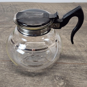 May include: A clear glass coffee carafe with a black lid and handle. The carafe has silver horizontal stripes and a silver rim. The handle is curved and attached to the side of the carafe. The lid has the word "Cole" printed on it.