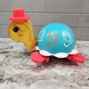 May include: A vintage pull-along toy turtle with a yellow head and a blue shell. The turtle has a red top hat, red feet, and a white scalloped edge around the shell. The shell features orange and white designs. A yellow string is attached for pulling.