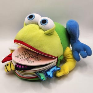 May include: A plush frog toy with large, googly eyes and a book in its mouth. The frog is primarily green and yellow, with blue and yellow limbs. The book's cover is pink with the title "Monday the Bullfrog."