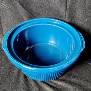 May include: A vibrant blue ceramic slow cooker insert. The insert has a round shape with a wide rim and two side handles. The exterior features vertical ribbed detailing, adding texture to the design. The interior is smooth and glossy.