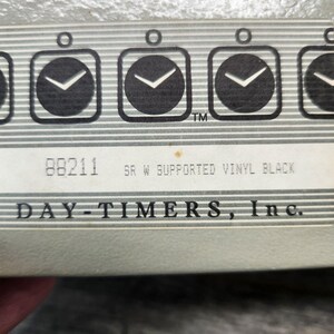 May include: A product label featuring a repeating pattern of black clock icons on a gray striped background. The text reads 88211 SR W SUPPORTED VINYL BLACK and DAY-TIMERS, Inc. The label is printed on a light gray surface with a horizontal orientation.