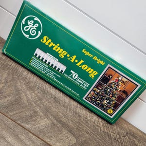 May include: A green rectangular box for a GE String-A-Long light set containing 70 clear miniature-style bulbs. The packaging features the GE logo, yellow text, and a photograph of a decorated Christmas tree. It lists special features.