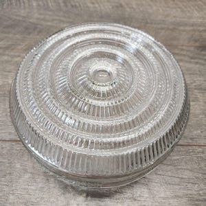 May include: Clear glass lid with a circular, ridged design. The lid features concentric rings of vertical lines, creating a textured surface. The glass is transparent, allowing light to pass through.