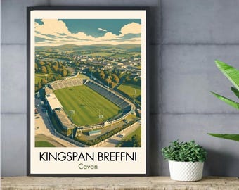 May include: A framed art print featuring an aerial illustration of Kingspan Breffni stadium in Cavan, Ireland. The image shows a green sports field surrounded by stadium seating, trees, and rolling hills under a blue sky with white clouds.