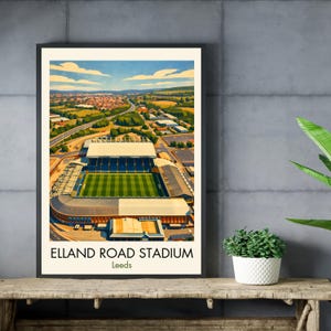 May include: A framed print of Elland Road Stadium in Leeds, featuring a detailed aerial view of the stadium with a green field and blue seating. The artwork showcases a vibrant landscape with a blue sky and surrounding buildings. The text "ELLAND ROAD STADIUM Leeds" is displayed at the bottom.