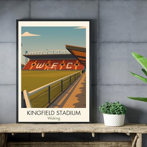 May include: A framed art print of Kingfield Stadium in Woking. The illustration features the stadium's red and orange facade with the letters "WFC" visible. A green field and blue sky complete the scene. The print is displayed on a wooden surface.