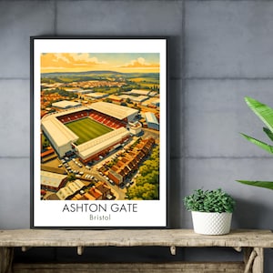 May include: A framed print of Ashton Gate stadium in Bristol, England. The artwork depicts the stadium with a green field, red seating, and white structures. The sky is a mix of yellow and blue. The text "ASHTON GATE Bristol" is at the bottom.
