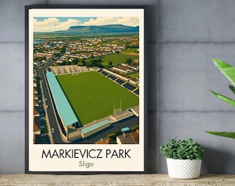 May include: A framed art print featuring an aerial illustration of Markievicz Park in Sligo, Ireland. The image displays a green sports field, a light blue roofed grandstand, surrounding residential buildings, and a distant mountain range under a blue sky.