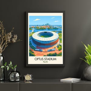 May include: A framed art print of Optus Stadium in Perth, Australia. The stadium is illustrated with a blue and white roof, surrounded by green fields and water. The text "OPTUS STADIUM Perth" is at the bottom of the print.