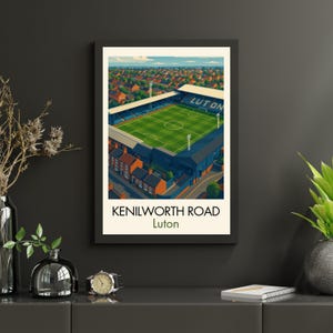 May include: A framed art print of Kenilworth Road stadium in Luton. The artwork depicts the stadium with a green field, blue seating, and the word "LUTON" on the roof. The print is set against a backdrop of red-roofed houses.