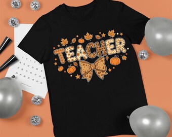 Puede incluir: Camiseta negra con la palabra "TEACHER" en un diseño otoñal. La palabra "TEACHER" está decorada con un estampado de cuadros y leopardo, rodeada de calabazas, hojas y un lazo.
