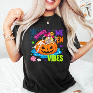 May include: A black t-shirt featuring a graphic of a skeleton wearing a pink hat and heart-shaped sunglasses, lounging in a pumpkin floatie on blue water. The text reads "SUMMER WEEN VIBES" in bright colors, surrounded by a spiderweb and seashells.
