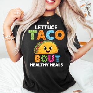Può includere: T-shirt nero con una grafica colorata che dice "Lettuce Taco Bout Healthy Meals". Il design presenta un taco sorridente e testo in vari colori, tra cui verde, arancione, blu e rosa.