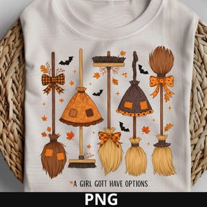 May include: White t-shirt featuring a whimsical design of five decorative broomsticks in shades of brown, orange, and tan, adorned with bows, leaves, and bats. The text "A GIRL GOTTA HAVE OPTIONS" is printed below the brooms. The PNG file format is also displayed.