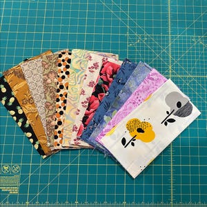 May include: A selection of fabric swatches in diverse patterns and colours, including floral, geometric, and abstract designs. The fabrics are arranged in a fan shape, showcasing the diverse textures and styles. The background is a green cutting mat.
