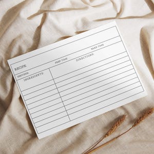 May include: A white recipe card with black text, including fields for recipe name, servings, ingredients, prep time, bake time, and directions. The card is on a beige fabric surface, with two sprigs of wheat in the bottom right corner.