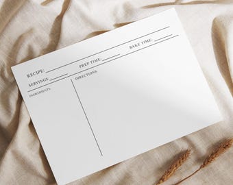 May include: A white recipe card with lines for recipe details, prep time, and bake time. The card includes spaces for ingredients, servings, and directions. The card is on a beige fabric surface, with wheat stalks in the bottom right corner.