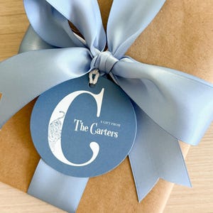 May include: A brown paper wrapped gift tied with a wide light blue satin ribbon bow. A round blue gift tag hangs from the ribbon, featuring a large white letter C with a tiger illustration inside and the text A GIFT FROM The Carters.