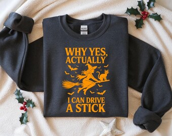 May include: Dark gray sweatshirt with an orange graphic of a witch on a broom, a cat, and bats. Text reads "WHY YES, ACTUALLY I CAN DRIVE A STICK". A cozy, long-sleeved garment for Halloween.