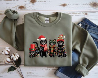 May include: An olive green sweatshirt featuring a graphic of four black cats dressed for Christmas. The cats wear Santa hats, scarves, and sunglasses, and are adorned with Christmas lights. The sweatshirt is paired with blue jeans.