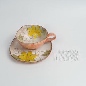 May include: A handmade ceramic teacup and saucer set with an irregular, organic shape. The exterior and interior feature a soft peach glaze with hand-painted yellow and white daisy-like flowers and green leaves. A small card with Japanese text is included.