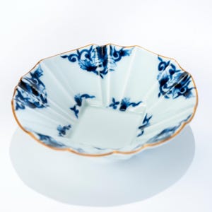以下が含まれることがあります： 白磁のボウル。縁は波型で、内側は正方形。青い花の模様と金の縁で装飾されています。デザインは対称的でエレガントです。