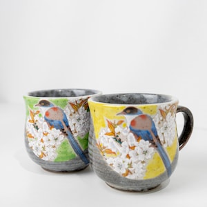 以下が含まれることがあります： 鳥と花の模様が施された2つのセラミックマグ。1つのマグは緑色の背景、もう1つは黄色です。各マグには、青、赤、茶色の羽を持つ鳥が白い花の中に止まっています。マグには灰色のリムとハンドルが付いています。
