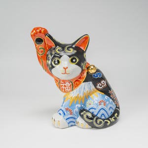 May include: A ceramic Maneki-neko cat figurine with a black, white, and orange coat. The cat has one raised paw, a gold bell around its neck, and a red collar featuring the kanji character for good fortune. The body is decorated with blue waves.