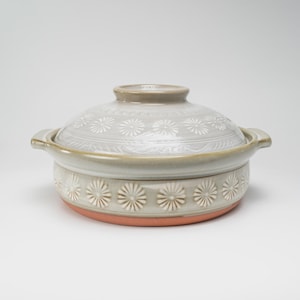 May include: A round ceramic lidded pot featuring a light grey glaze with a repeating embossed white daisy pattern around the lid and base. The pot has two side handles and a visible terracotta-coloured clay bottom edge with a smooth finish.