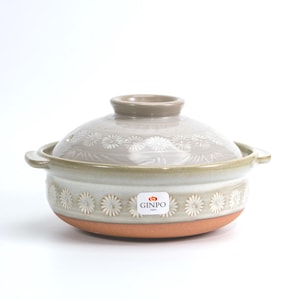 Puede incluir: Una olla japonesa donabe de cerámica gris claro con tapa a juego. La olla presenta un patrón floral alrededor del borde y en la tapa. La base es de color marrón rojizo. La marca "GINPO Japan" está en una etiqueta.