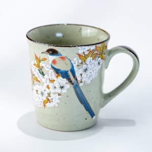 May include: A ceramic mug with a pale green speckled finish. The mug features a detailed illustration of a blue and white bird perched on a branch with white flowers and yellow leaves. The rim and handle are a darker green.