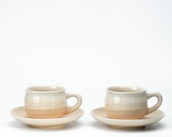 May include: Two ceramic cups and matching saucers featuring a smooth, rounded shape. Each piece has a two-tone glaze, with a creamy off-white finish on the upper portion that transitions into a soft, warm peach hue at the base.