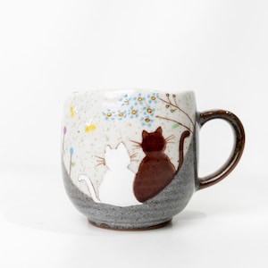 以下が含まれることがあります： 茶色の持ち手と猫のデザインが施されたセラミックマグ。白い猫と茶色の猫が一緒に座っており、青い花のアクセントとまだらの背景が特徴です。マグカップは薄いベージュ色です。