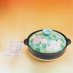 Pode incluir: Uma panela donabe japonesa de cerâmica com um design floral. A tampa apresenta um fundo verde claro com flores rosa, brancas e roxas. A panela tem uma borda e alças pretas, com uma base branca.