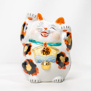 Peut inclure: Figurine Maneki Neko en céramique blanche avec des taches orange et noires, un collier bleu et une cloche dorée. Le chat sourit et lève une patte. Une plaque dorée avec des caractères japonais est suspendue à son cou.