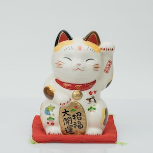 Puede incluir: Una figura de gato maneki-neko de cerámica blanca con manchas negras y naranjas, que lleva un collar rojo con un cascabel dorado. El gato tiene una pata levantada y sostiene una moneda dorada con los caracteres japoneses negros 大開運 y 招福. Otros motivos pintados incluyen una tortuga, una grulla y un pino.