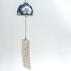 Op de afbeelding: Een keramische windgong met een donkerblauw en wit bloemenpatroon. De windgong heeft een klokvormige bovenkant met witte bloempatronen en een hangend label met Japanse tekst. De gong hangt aan een zwarte draad.