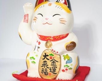 Puede incluir: Una figura de gato Maneki-neko de cerámica blanca con marcas negras, naranjas y doradas. El gato tiene la pata derecha levantada, un collar rojo con un cascabel dorado y una barriga ovalada dorada con caracteres kanji negros y pinos.